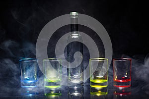 Five multicolored empty shot glasses and small bottle placed symmetrically on a black background in a theatrical smoke