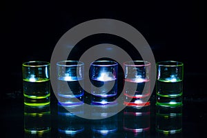 Five multicolored full of drinks shot glasses symmetrically placed a black background.