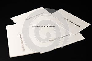 Five notes with words `Quality Guaranteed` are on the black table/background.  World Quality Day