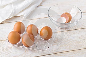 Five raw chicken eggs in a transparent plastic tray and empty eggshell in a glass bowl on a white wooden table. Cooking for easter