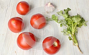 Five red large tomatoes are next to a bunch of greens and a head of garlic on a light background