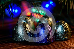 Five sweets wrapped in foil lie on a wooden surface. Background - Christmas tree and multi-colored LED lights. Night magic. Bokeh