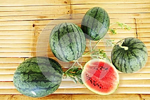 Five watermelon with a slice and leaves on a bamboo table.