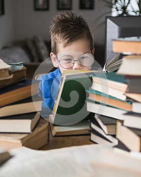 Five year old boy in glasses reading a book with a stack of books next to him