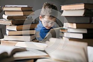 Five year old boy in glasses reading a book with a stack of books next to him