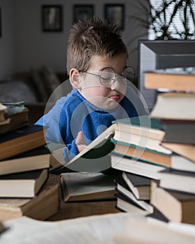 Five year old boy in glasses reading a book with a stack of books next to him. Smart intelligent preschool kid choosing books to