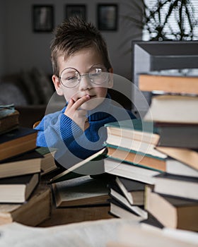 Five year old boy in glasses reading a book with a stack of books next to him. Smart intelligent preschool kid choosing books to