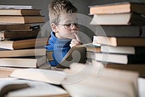 Five year old boy in glasses reading a book with a stack of books next to him. Smart intelligent preschool kid choosing books to