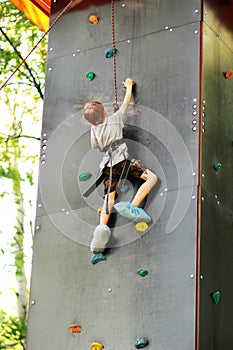 Five year old boy learning to climb the rock wall outside in the summer park.