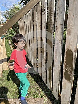 A five-year-old cute curly-haired black-haired boy in green pants and a red T-shirt looks like a painted wooden fence. The concept