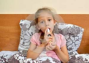 A five-year-old girl holds a spray in her hand and irrigates their throat while lying in bed. Self-treatment photo