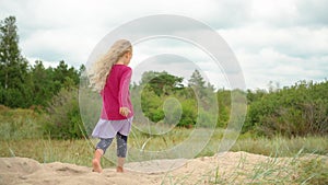Five years old blonde girl with pink shirt walking barefoot on top of dune sand