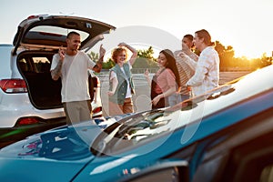 Five young well-dressed friends dancing to music and smiling together outside on a parking site near their cars with a