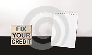Fix Your Credit text written on building blocks and white and black background, next to a notepad for writing.Conceptual photo.