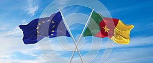 flags of European Union and Cameroon waving in the wind on flagpoles against sky with clouds on sunny day. Symbolizing