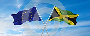 flags of European Union and Jamaica waving in the wind on flagpoles against sky with clouds on sunny day. Symbolizing