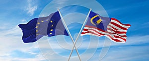 flags of European Union and Malaysia waving in the wind on flagpoles against sky with clouds on sunny day. Symbolizing
