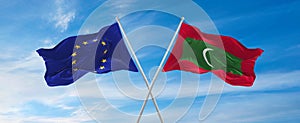 flags of European Union and Maldives waving in the wind on flagpoles against sky with clouds on sunny day. Symbolizing