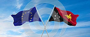 flags of European Union and Papua New Guinea waving in the wind on flagpoles against sky with clouds on sunny day. Symbolizing