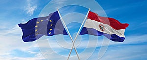 flags of European Union and Paraguay waving in the wind on flagpoles against sky with clouds on sunny day. Symbolizing