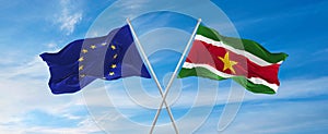 flags of European Union and Suriname waving in the wind on flagpoles against sky with clouds on sunny day. Symbolizing