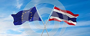 flags of European Union and thailand waving in the wind on flagpoles against sky with clouds on sunny day. Symbolizing