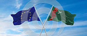 flags of European Union and Turkmenistan waving in the wind on flagpoles against sky with clouds on sunny day. Symbolizing