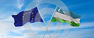 flags of European Union and Uzbekistan waving in the wind on flagpoles against sky with clouds on sunny day. Symbolizing