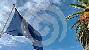 Flags of the European Union waving in the wind at sunny day, blue sky with clouds on background, palm trees