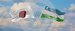 flags of Japan and Uzbekistan waving in the wind on flagpoles against sky with clouds on sunny day. Symbolizing relationship,