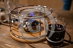 Transparent glass mug with hot mulled wine inside near metal burner for traditional French cheese fondue. Blue flaming fire