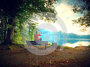 Flare, soft focus. Man sit on wooden bench at mountain lake . Bank under beeches tree