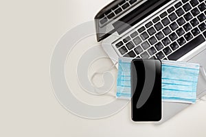 Flat composition of black and white laptop keyboard, phone, coffee cup and blue medical mask on wooden table background. Top view