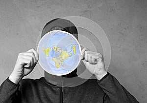 Flat Earther concept. Person who believes that Earth is flat disc. Anonymous hooded Man holding flat Earth model in front of face.