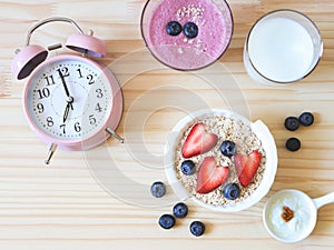 flat lay of alarm clock, breakfast with oat or granola in white bowl, fresh blueberries, strawberries, a glass of milk, blueberry