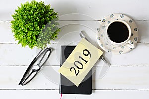 Flat lay of artificial green plant, cup of coffee, eye glass,,note book, pen and paper with number 2019 on white wooden pallet