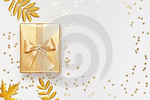 Flat lay Autumn holiday concept. Golden gift or present box with ribbon, confetti stars, golden autumn leaves on light gray