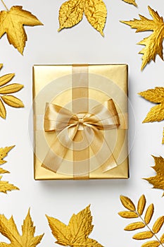 Flat lay Autumn holiday concept. Golden gift or present box with ribbon and golden autumn leaves on light gray background top view