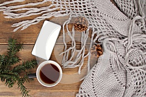 Flat lay background with a mug of tea, white smart phone with blank screen, shawl with fringe, coniferous branches and dried