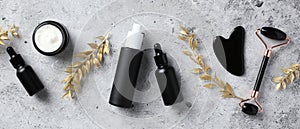 Flat lay black obsidian face roller, gua sha, serum, essential oils and golden branches on stone table. Luxury skincare cosmetics