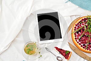 Flat lay breakfast in bed with raspberry cheesecake, mint tea and open note book, tablet