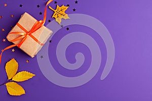 Flat lay of brown gift box with a orange satin ribbon bow and stars shapes yellow leaves on vibrant purple background with copy