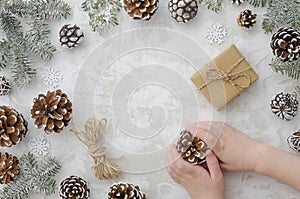 Flat lay children`s hands hold cone, craft handmade gift box, rope, fir branches and snowflakes on white background