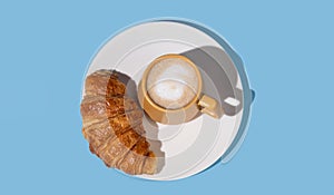 Flat lay of coffee cup and fresh croissant on a white plate on a blue background