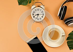 Flat lay  of coffee cup , headphones , alarmclock and mobile phone on orange background with monstera leaves decoration and copy