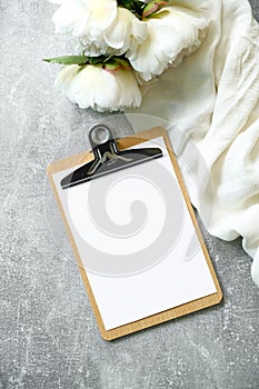 Flat lay composition with blank paper clipboard mockup, peony flowers and beige cloth on stone background. Top view wedding