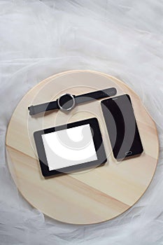 Flat lay composition with a blank tablet, blank mobile phone a black wristwatch laid on a circular wooden table on a white tulle b