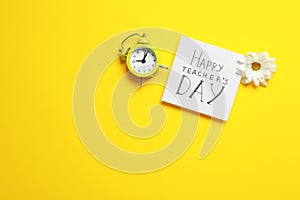 Flat lay composition of card with inscription HAPPY TEACHER`S DAY, flower and alarm clock on yellow background