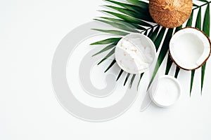Flat lay composition with coconut oil body cream, coconut half, grated coconut in bowl and tropical palm leaf on white background