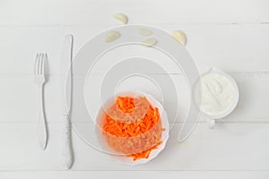 Flat lay, composition, freshly grated carrot in a plate and garlic. On the wooden table, a knife and fork. White